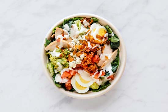 Aerial view of a salad bowl that has green veggies, chicken, gorgonzola, eggs, tomatoes, bacon, mashed avocado, and ranch dressing