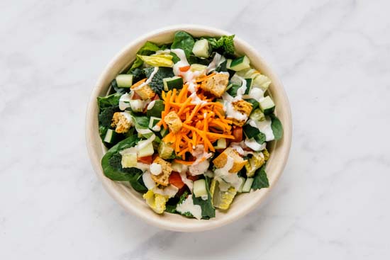 Aerial view of a salad bowl that has green veggies, pickled carrots, tomatoes, cucumbers, croutons, and ranch dressing