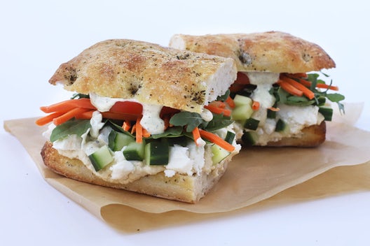 A sandwich cut in half that has kale, cucumber, hummus, feta cheese, pickled carrots, tomatoes, and tzatziki sauce on focaccia bread.