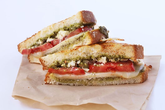 A sandwich cut in half stacked on top of one another that has white three different cheeses, tomato, and pesto on grilled cheesy bread.