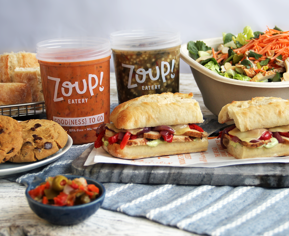 A table spread of bread, cookies, sandwiches, salad, and soup in a container that says Zoup!