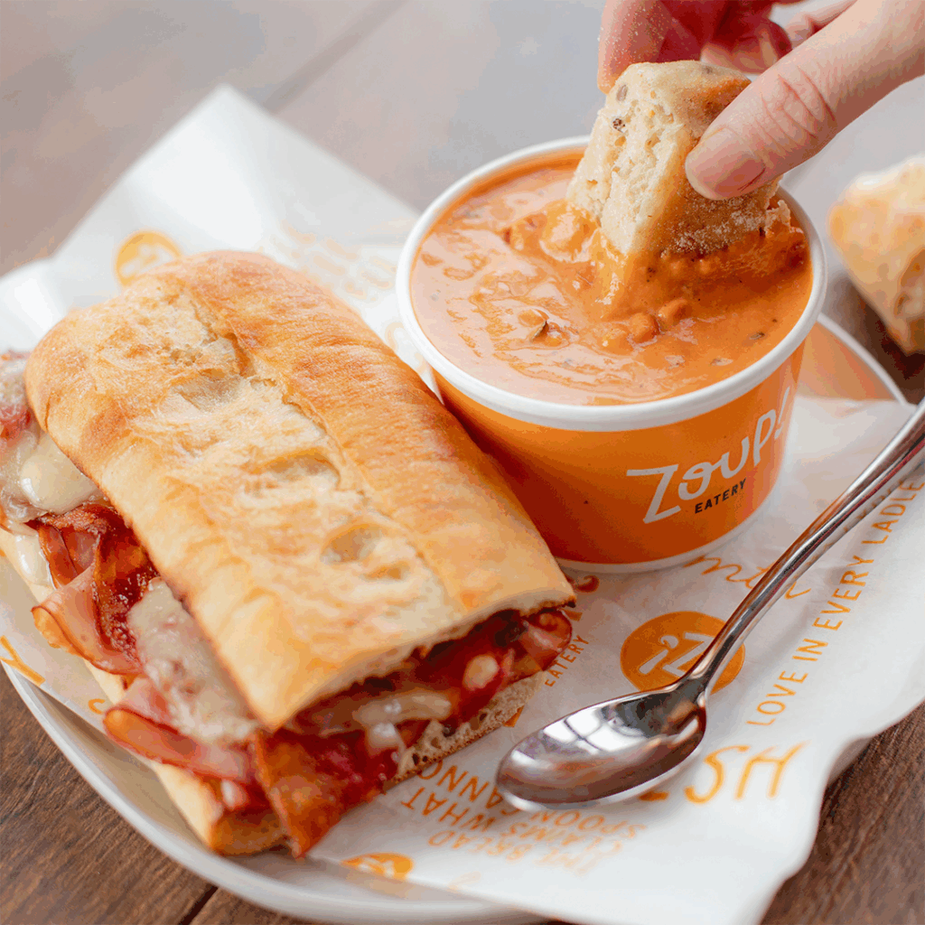 Toasted sandwich with melted cheese and meat beside a cup of creamy tomato soup, with a hand dipping bread into the soup. Zoup! Eatery branding is visible on the cup and wrapper.