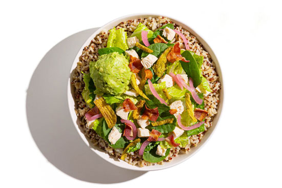 Southwest-style bowl with grilled chicken, corn, shredded cheese, tortilla strips, fresh cilantro, and a lime wedge over greens and grains.