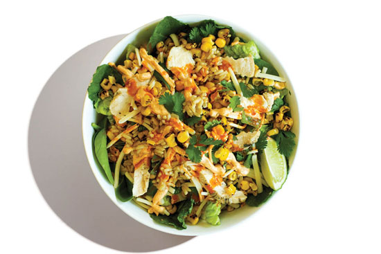 Southwest-style bowl with grilled chicken, corn, shredded cheese, tortilla strips, fresh cilantro, and a lime wedge over greens and grains.