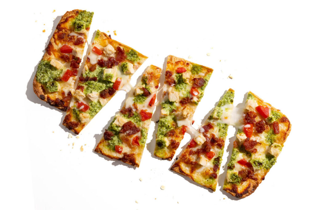 Sliced pesto chicken flatbread pizza with bacon bits, red bell peppers, and melted cheese.