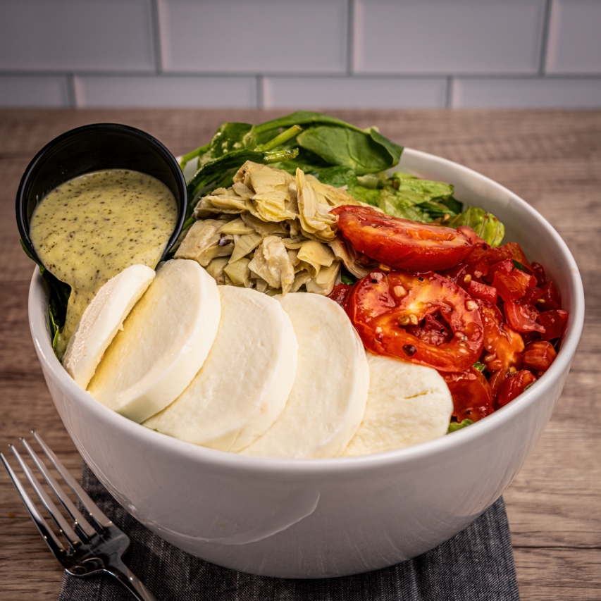 A fresh salad bowl with sliced mozzarella, artichoke hearts, tomatoes, leafy greens, and a cup of creamy herb dressing.