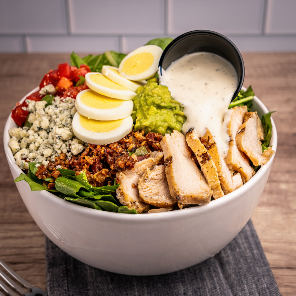 A colorful salad bowl topped with sliced grilled chicken, hard-boiled eggs, guacamole, blue cheese crumbles, bacon bits, diced tomatoes, and a cup of creamy dressing.