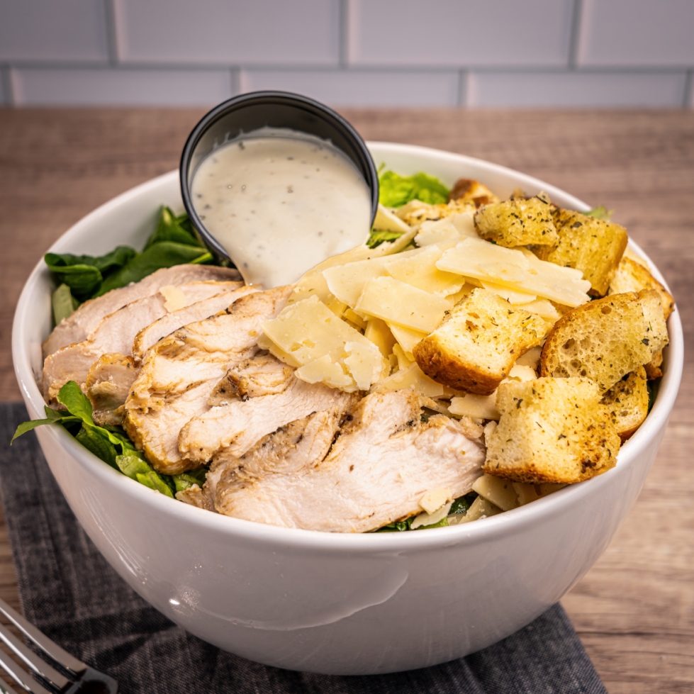 A classic chicken Caesar salad bowl with grilled chicken slices, shaved parmesan, croutons, and a cup of creamy Caesar dressing.