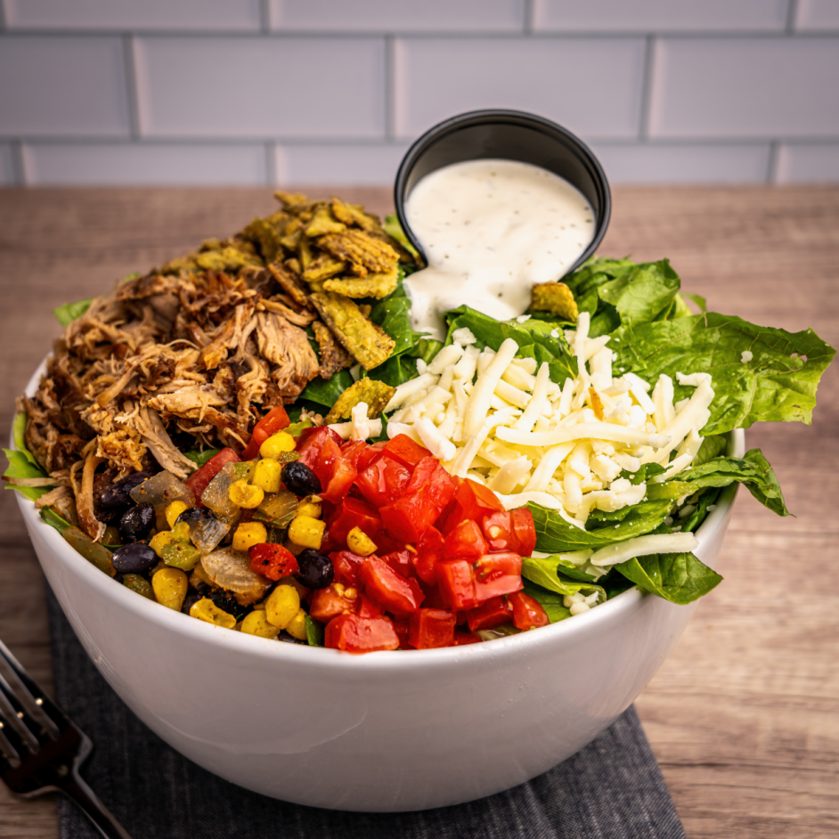 A colorful salad bowl topped with sliced grilled chicken, hard-boiled eggs, guacamole, blue cheese crumbles, bacon bits, diced tomatoes, and a cup of creamy dressing.
