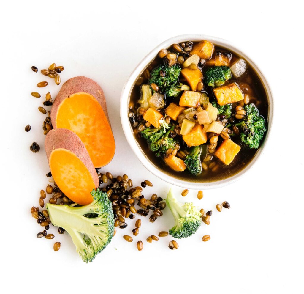 Bowl of vegetable stew with sweet potato, broccoli, onions, and grains in broth, surrounded by raw ingredients on a white background.