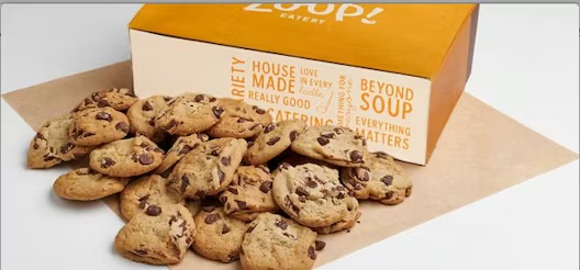 A pile of chocolate chip cookies in front of an orange and white Zoup! Eatery box on a sheet of parchment paper.