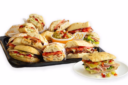 A catering tray filled with assorted ciabatta sandwiches containing deli meats and vegetables, with a small cup of relish in the center and one sandwich served separately on a white plate.