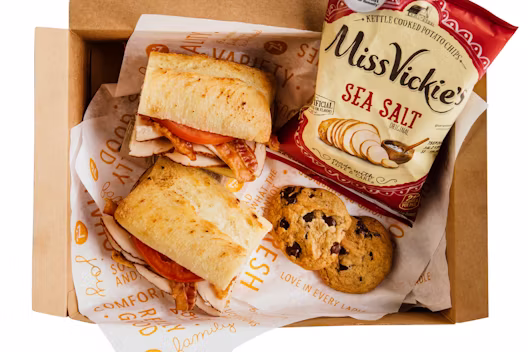 A boxed meal containing a ciabatta sandwich with sliced meat and tomato, two chocolate chip cookies, and a bag of Miss Vickie’s Sea Salt chips.