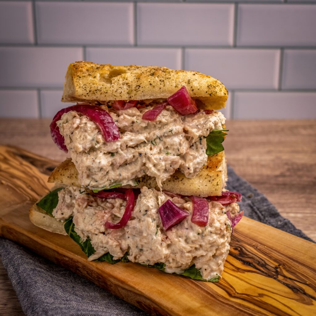 A stacked sandwich made with herbed focaccia bread, filled with creamy chicken salad, leafy greens, and pickled red onions.