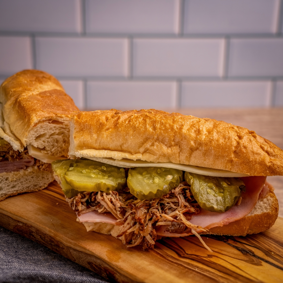 A close-up of a pulled pork and ham sandwich with pickles and cheese on a toasted baguette, resting on a wooden board.