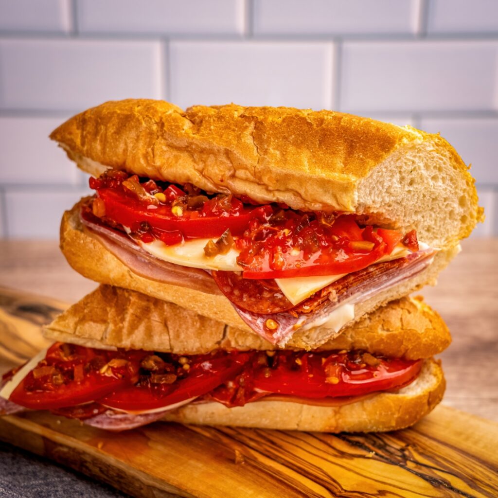 A stacked Italian sub sandwich filled with sliced tomatoes, deli meats, cheese, and chopped peppers on a toasted baguette.