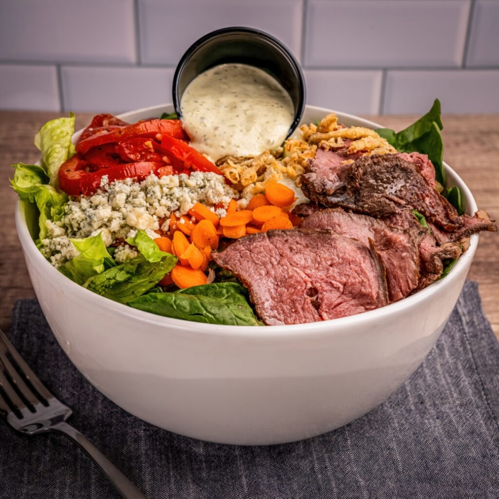 A hearty salad bowl with sliced steak, shredded carrots, roasted red peppers, blue cheese crumbles, crispy onions, and a cup of creamy dressing.