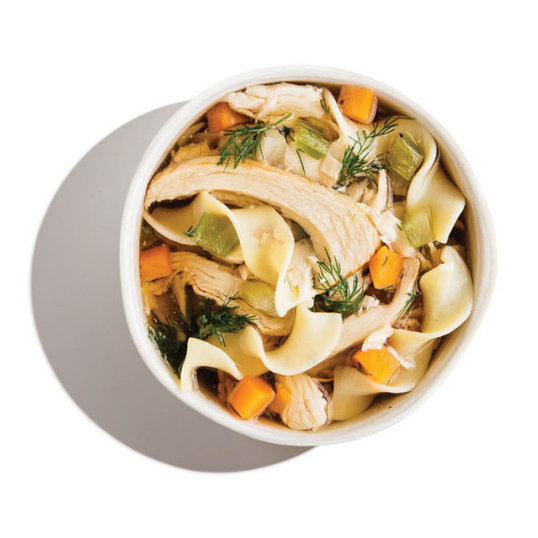 Overhead view of a white bowl of chicken noodle soup with wide egg noodles, shredded chicken, diced carrots, celery, and dill.