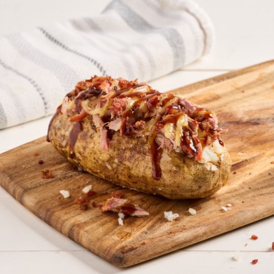 Loaded baked potato topped with bacon, melted cheese, and barbecue sauce on a wooden board.
