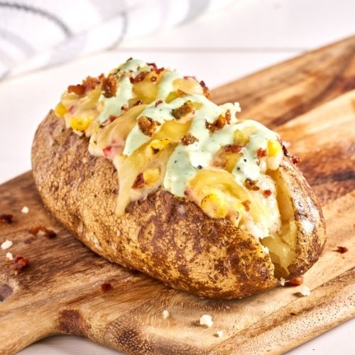 Baked potato topped with melted cheese, corn, bacon bits, and creamy herb sauce on a wooden board.