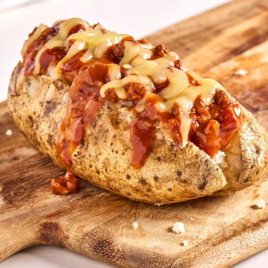 Chili cheese baked potato topped with red chili sauce, melted cheese, and bacon crumbles on a wooden board.